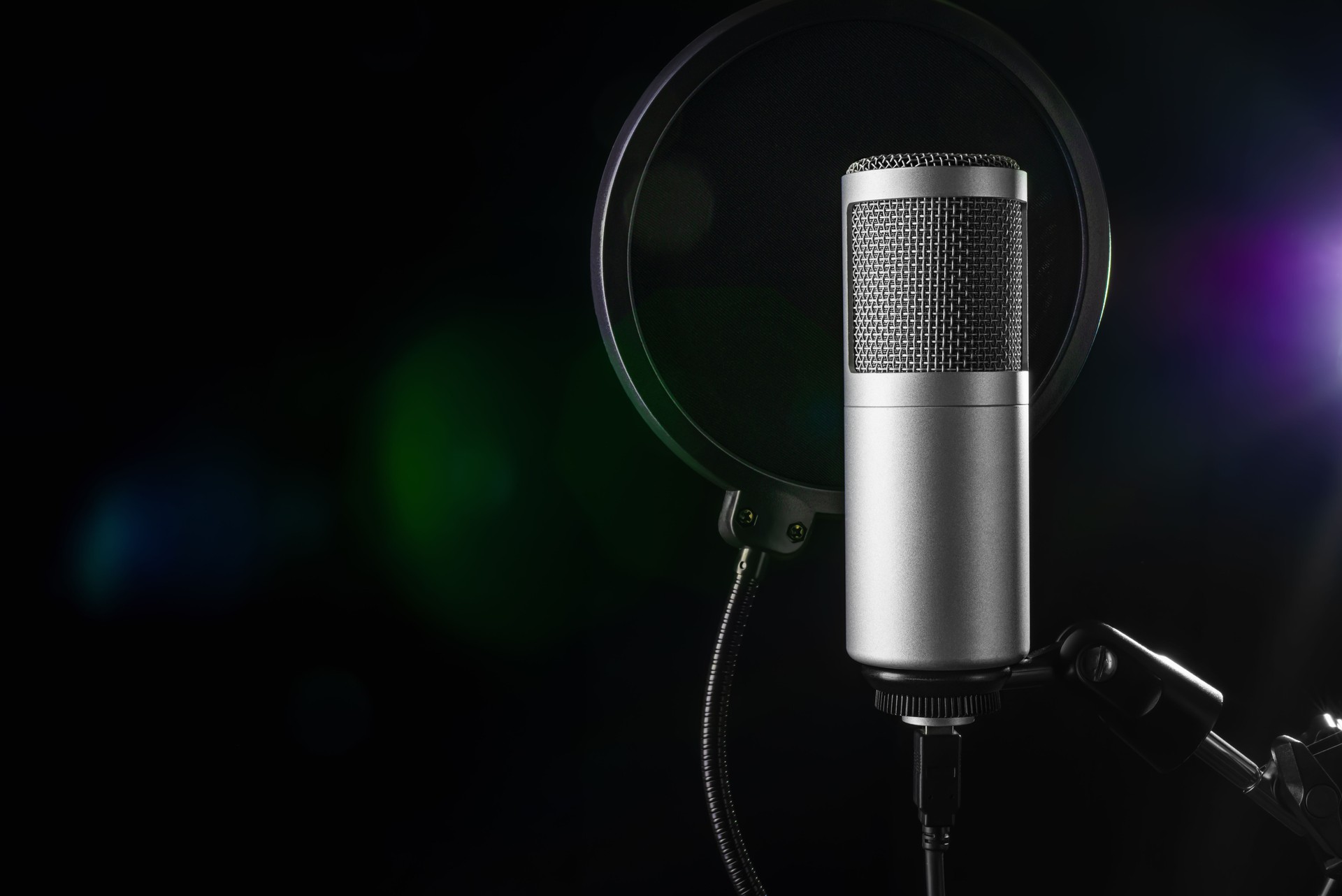 Studio microphone with a pop filter, poised for an audio recording session, set against a dark backdrop illuminated by colorful lights.